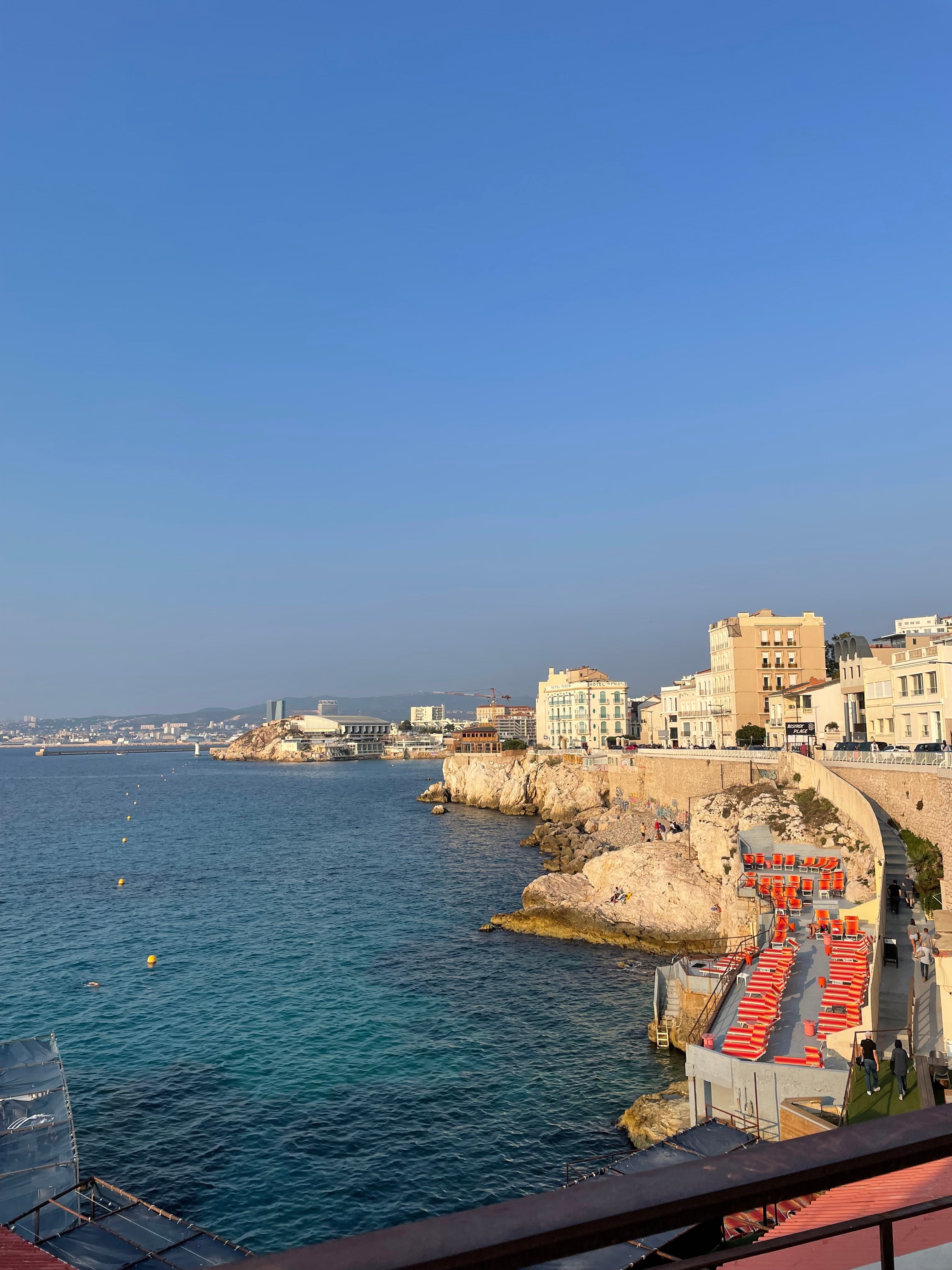 Marseille coastline with rocky shore, blue Mediterranean water, waterfront buildings, and red-striped lounge chairs