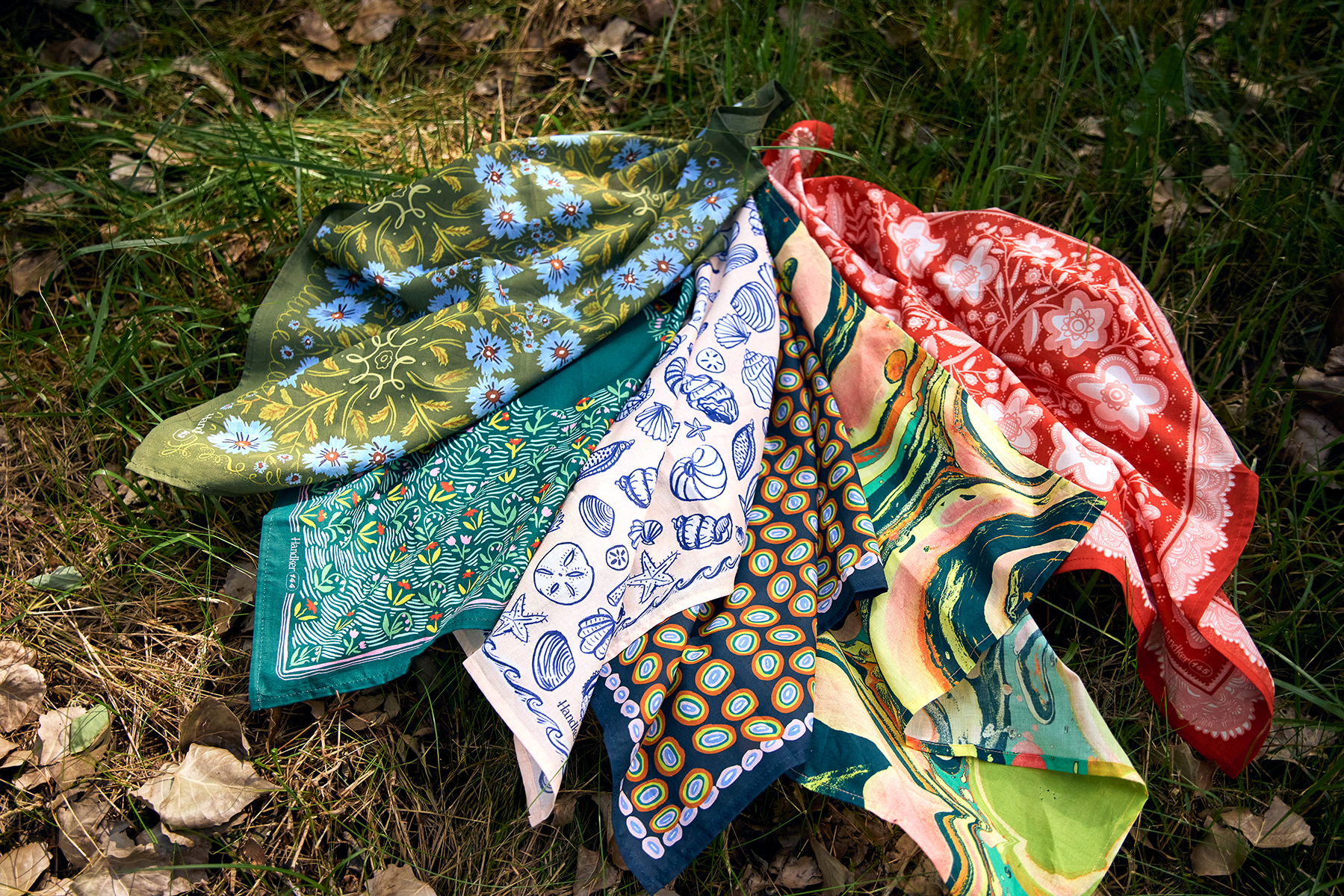 Spread of patterned bandanas laid on grass, featuring a pale pink bandana with blue seashells among floral and abstract prints