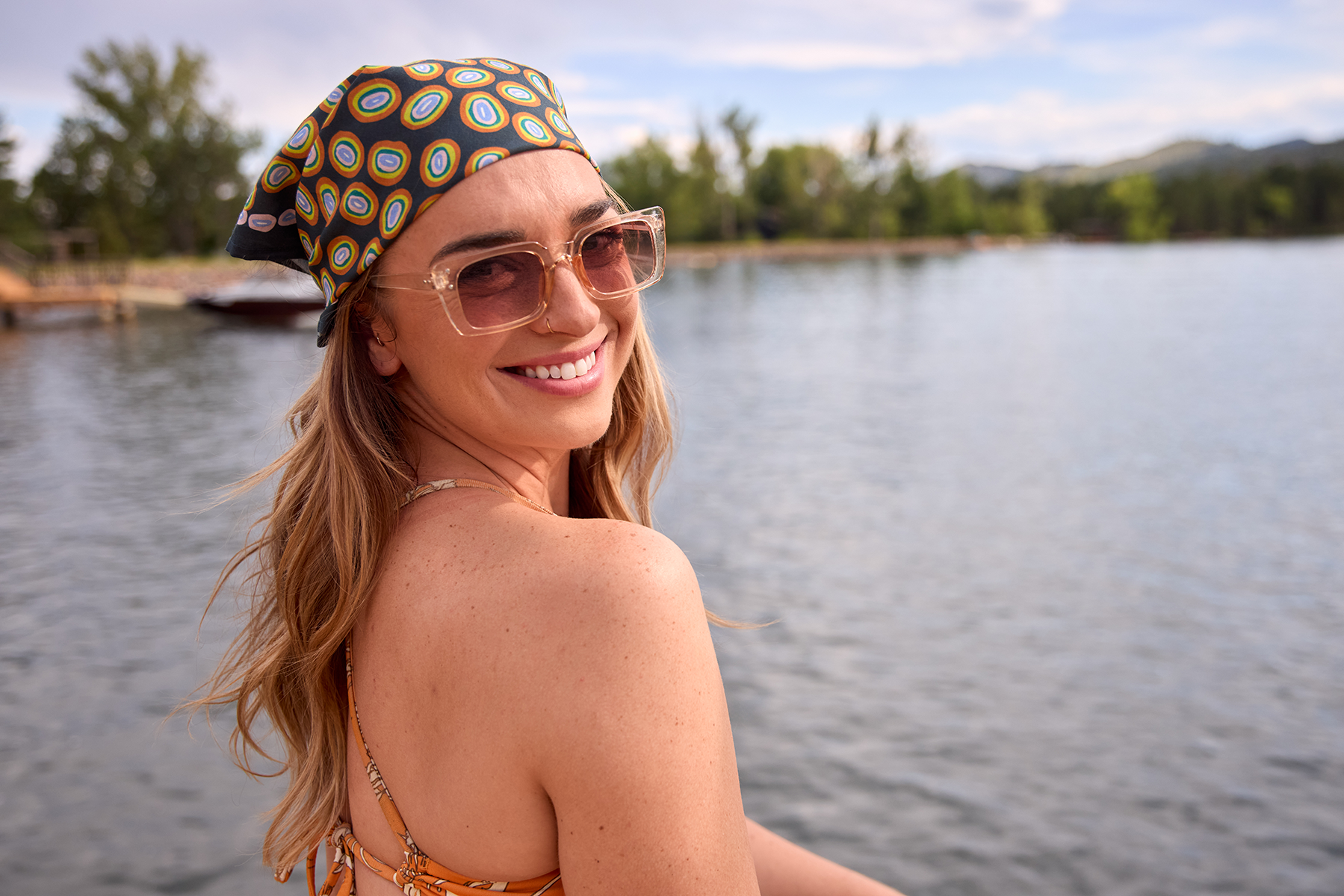 Smiling woman wearing a navy bandana with orange, gold, and pink circular pattern at a sunny lakeside