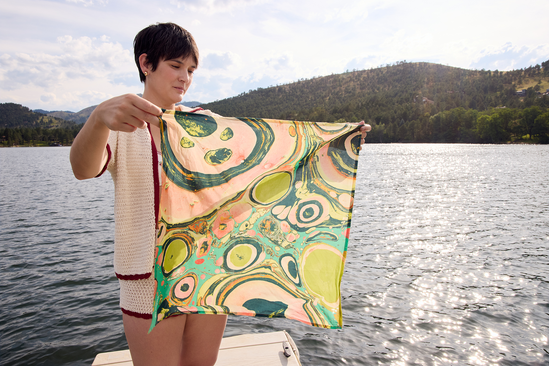 Front view of No. 140 Marble Bandana showing multicolored green and pink marble pattern held by a person on a dock with a sunlit lake and hills in the background
