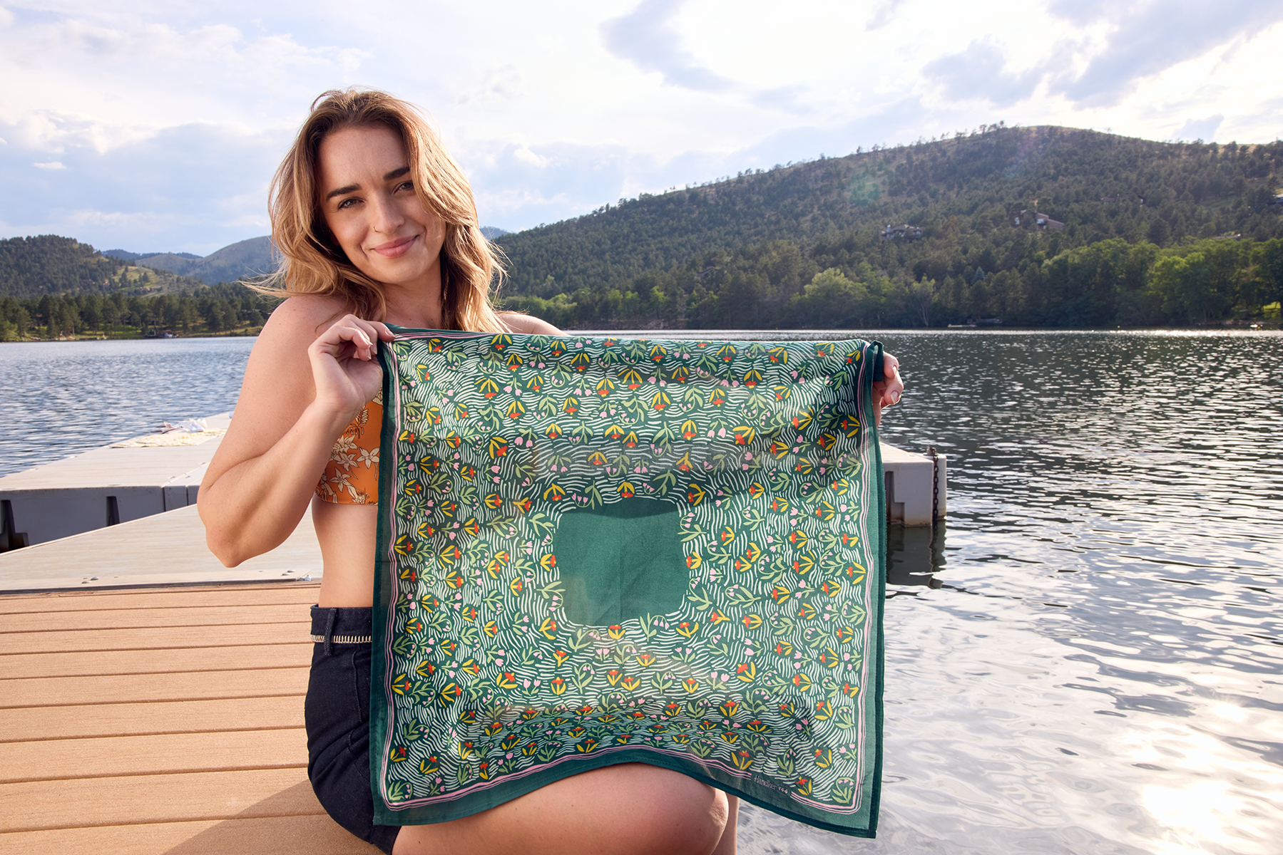 Woman sitting on a dock holding a 22 by 22 inch forest green floral bandana, lake and tree-covered hills in the background