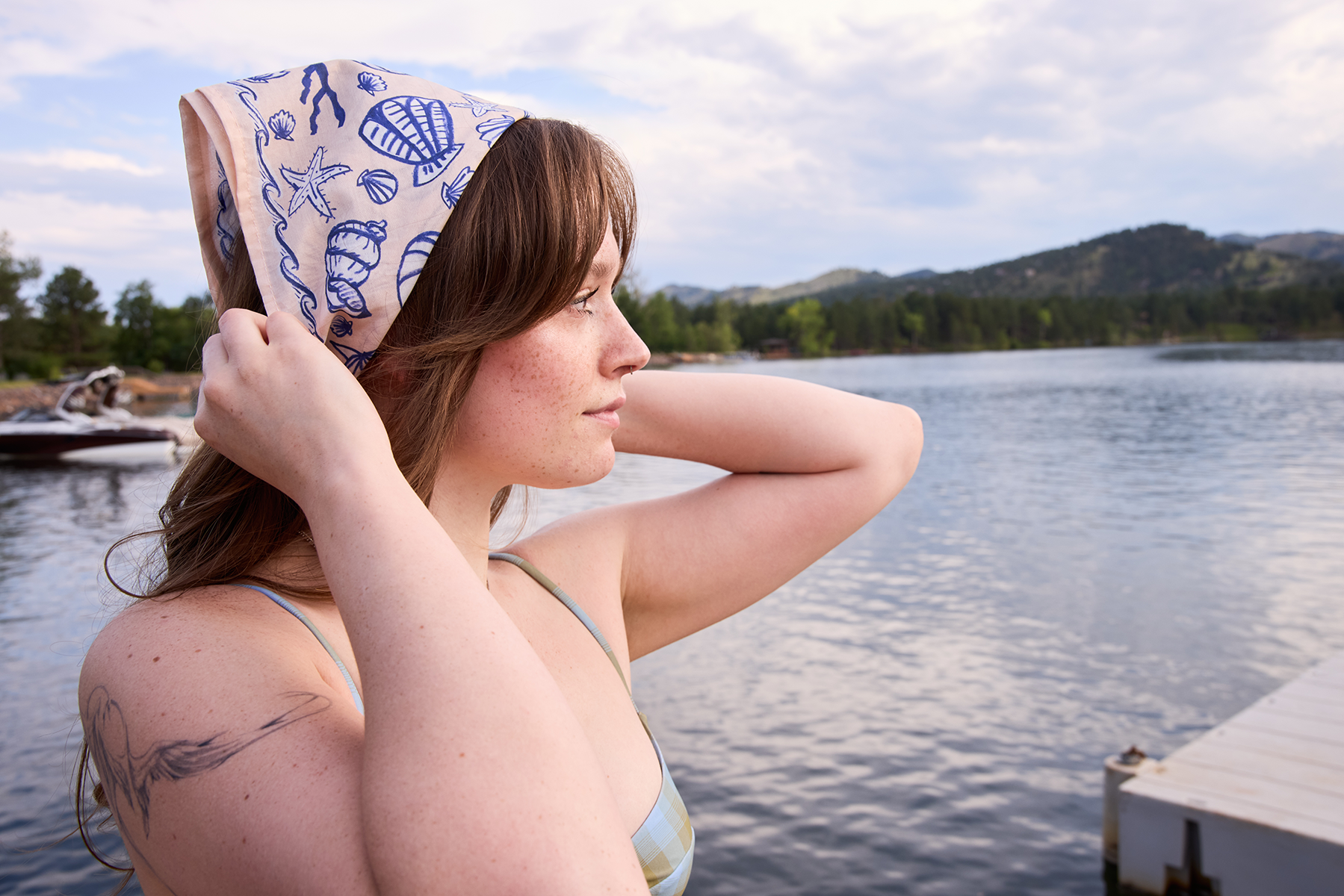 Side view of Seashore bandana worn as a headscarf showcasing pale peachy pink fabric with bright blue shell print at a lakeside dock