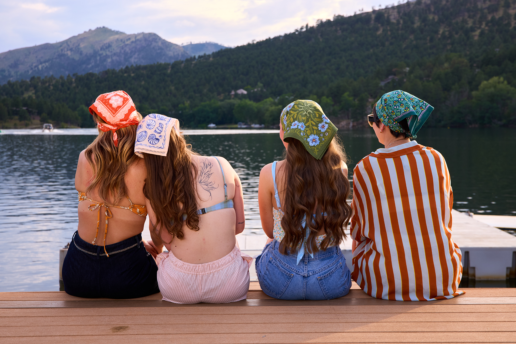 Four people wearing patterned bandanas sit on a wooden dock by a lake with tree-covered mountains in the background
