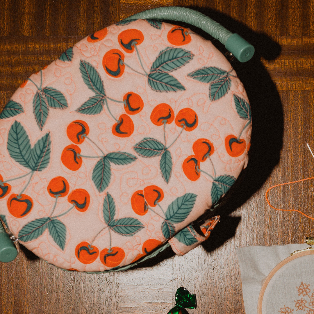 Round cherry-print fabric pouch resting on a wooden table next to an embroidery hoop, orange thread, and a green cord