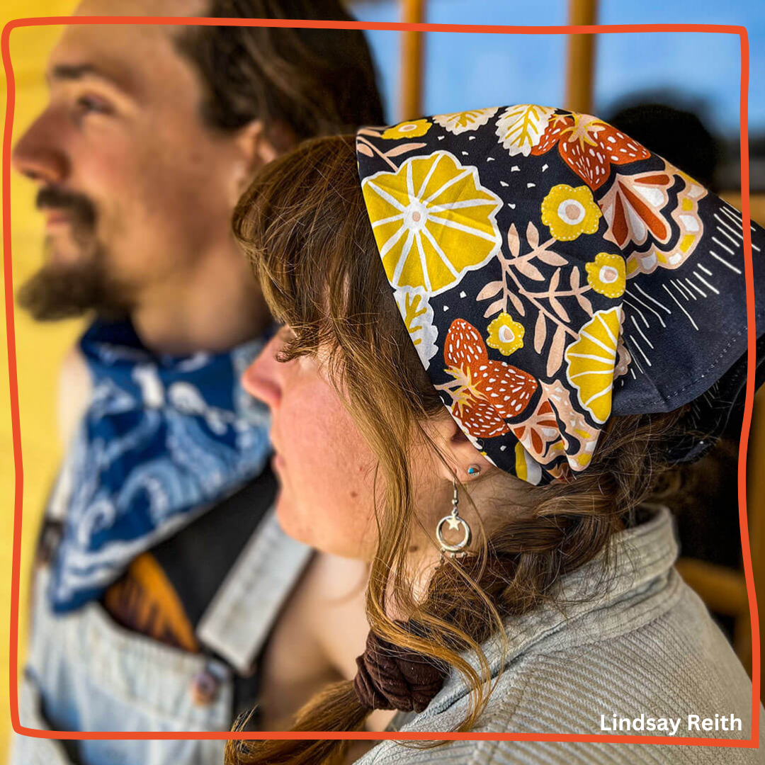Woman wearing a floral black, yellow, and orange bandana tied at the back, with a man blurred behind wearing a blue bandana