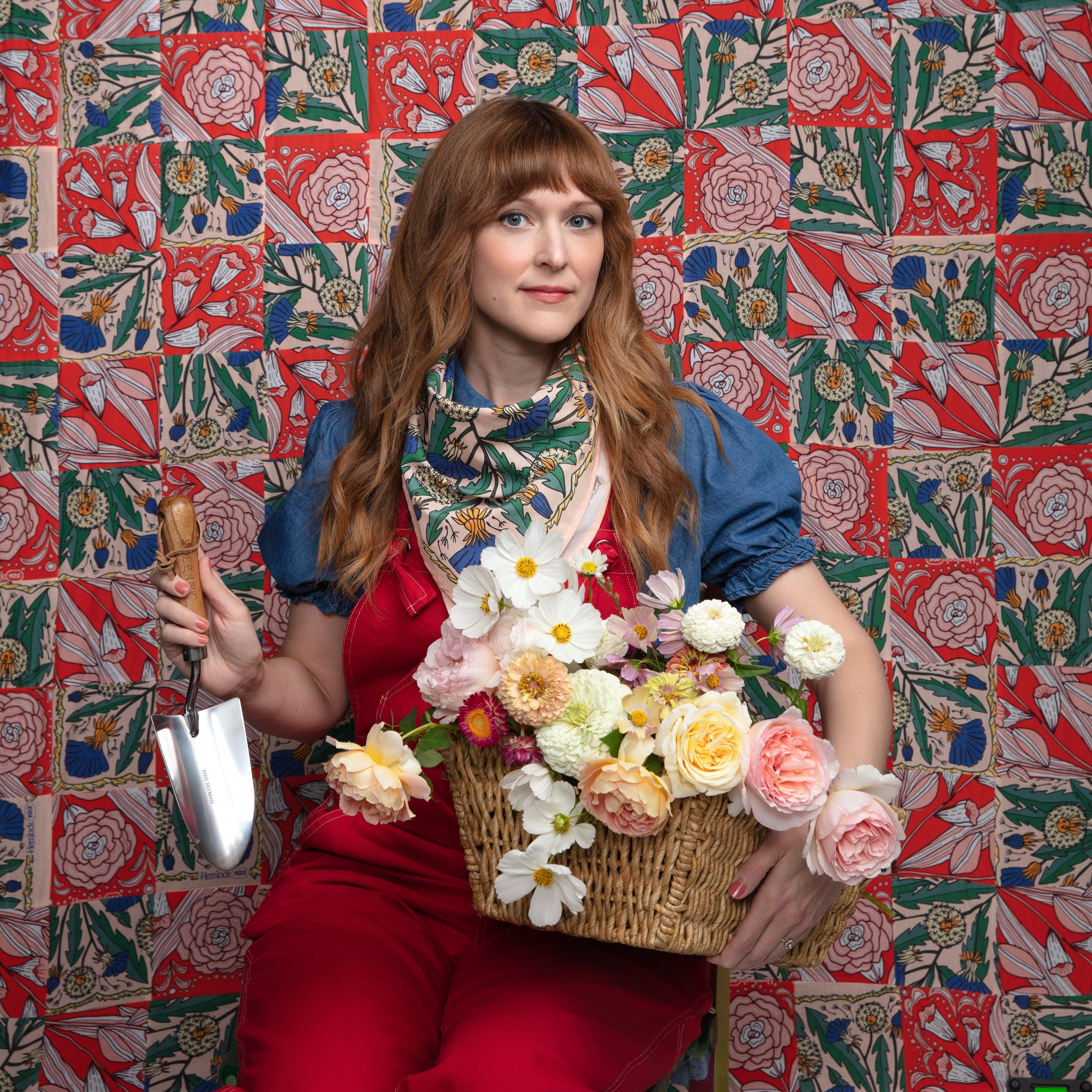 Woman wearing Kelly green floral bandana tied at neck, holding garden trowel and basket of flowers against matching backdrop