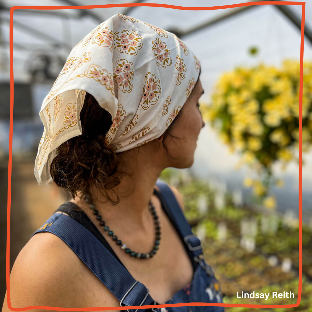 Woman wearing a cream Handker bandana with peach floral print tied over a low bun in a greenhouse with yellow flowers