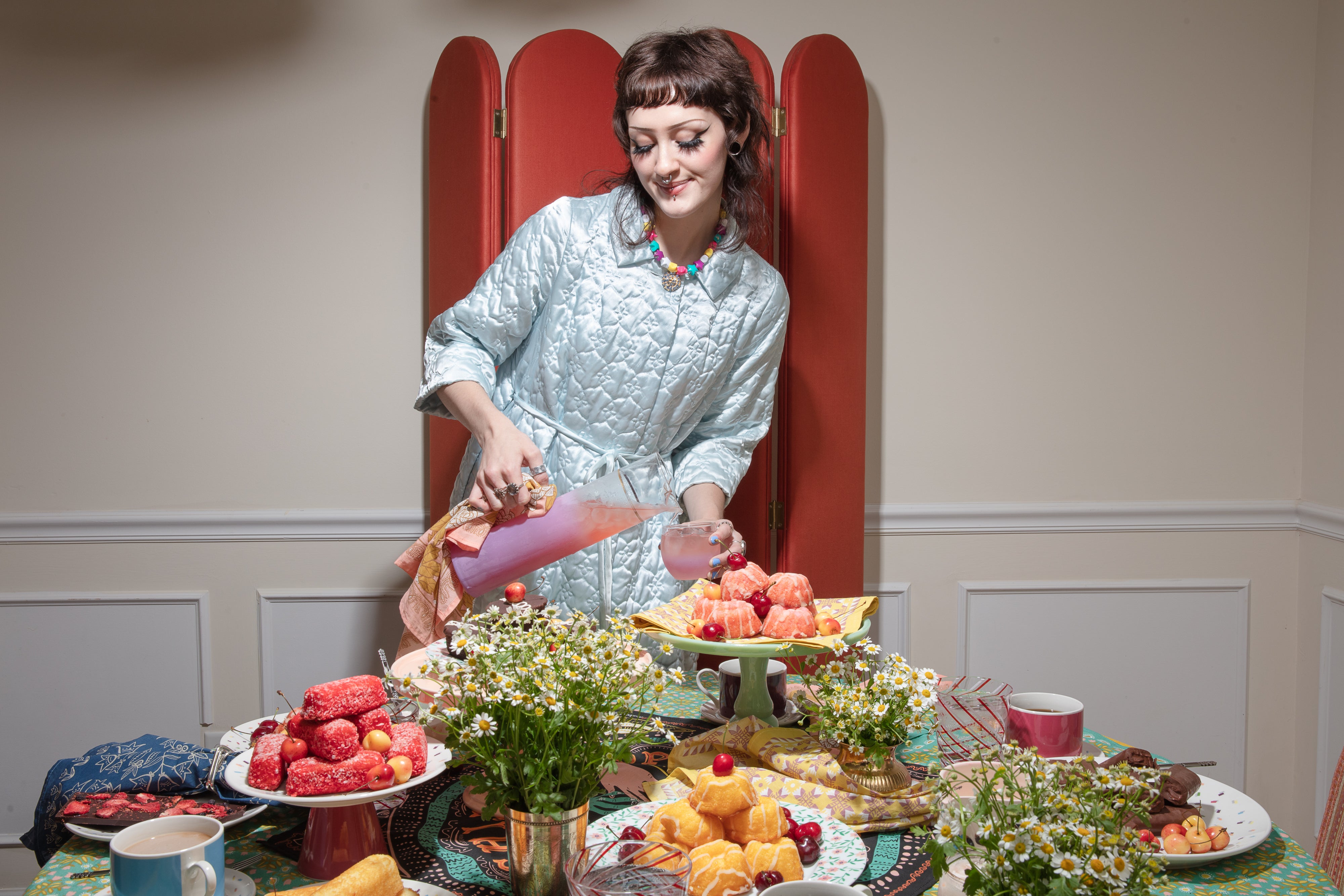 Woman pouring pink punch at a bandana-covered table set with pastries, fresh chamomile flowers, and folded bandana napkins