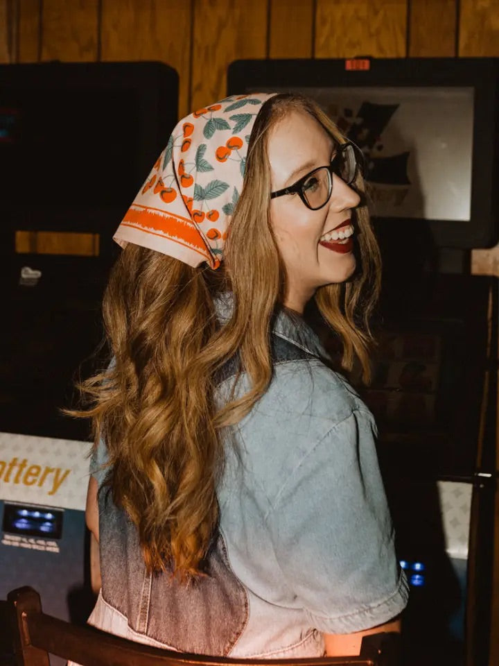 Three-quarter view of No 077 Cherry bandana on a woman showing peachy-pink fabric with red cherries and teal stems tied at the back