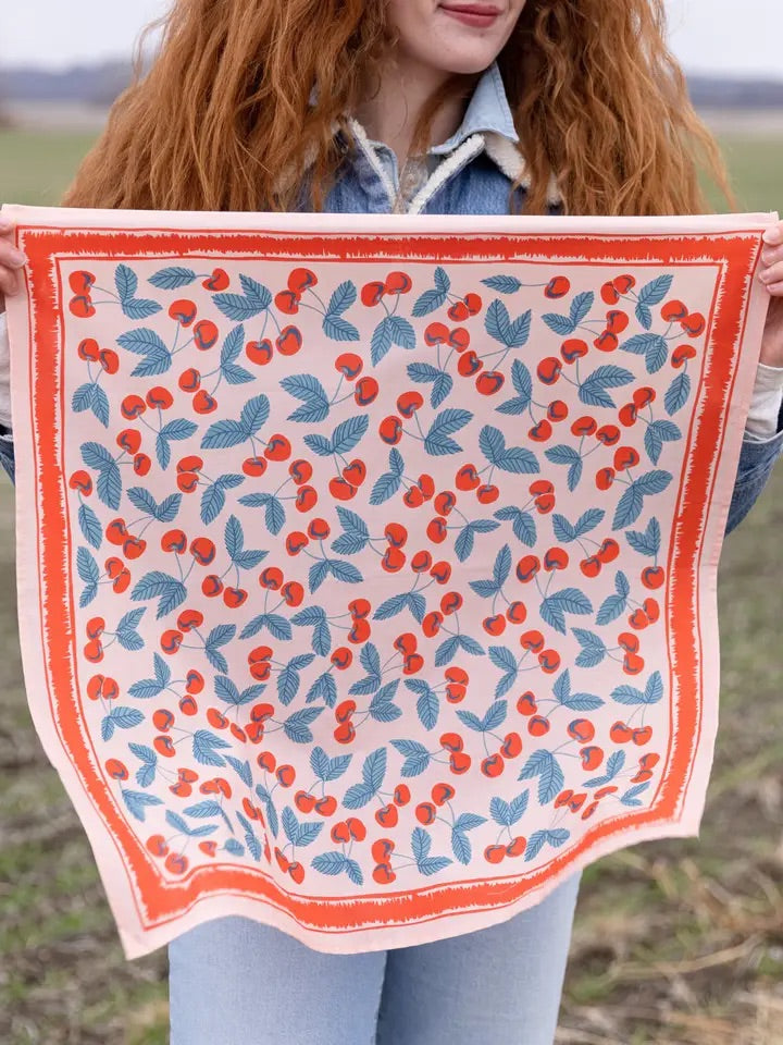 Front view of No. 077 Cherry bandana showing peachy-pink background with red cherries and teal leaves held by person outdoors