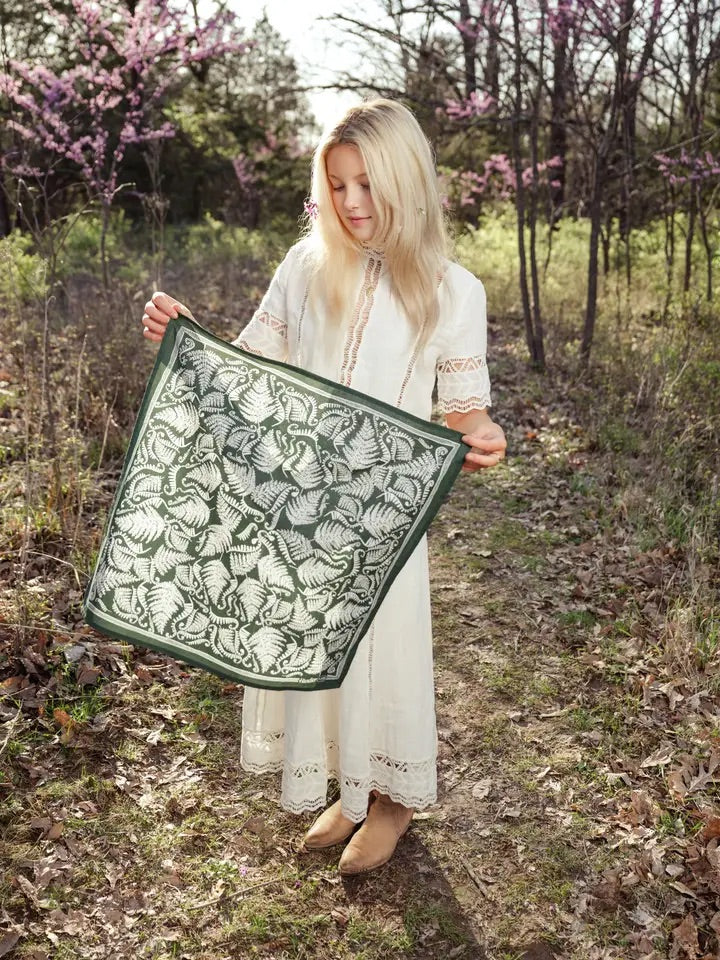 Young woman holding a dark forest green bandana printed with white ferns in a sunlit woodland clearing
