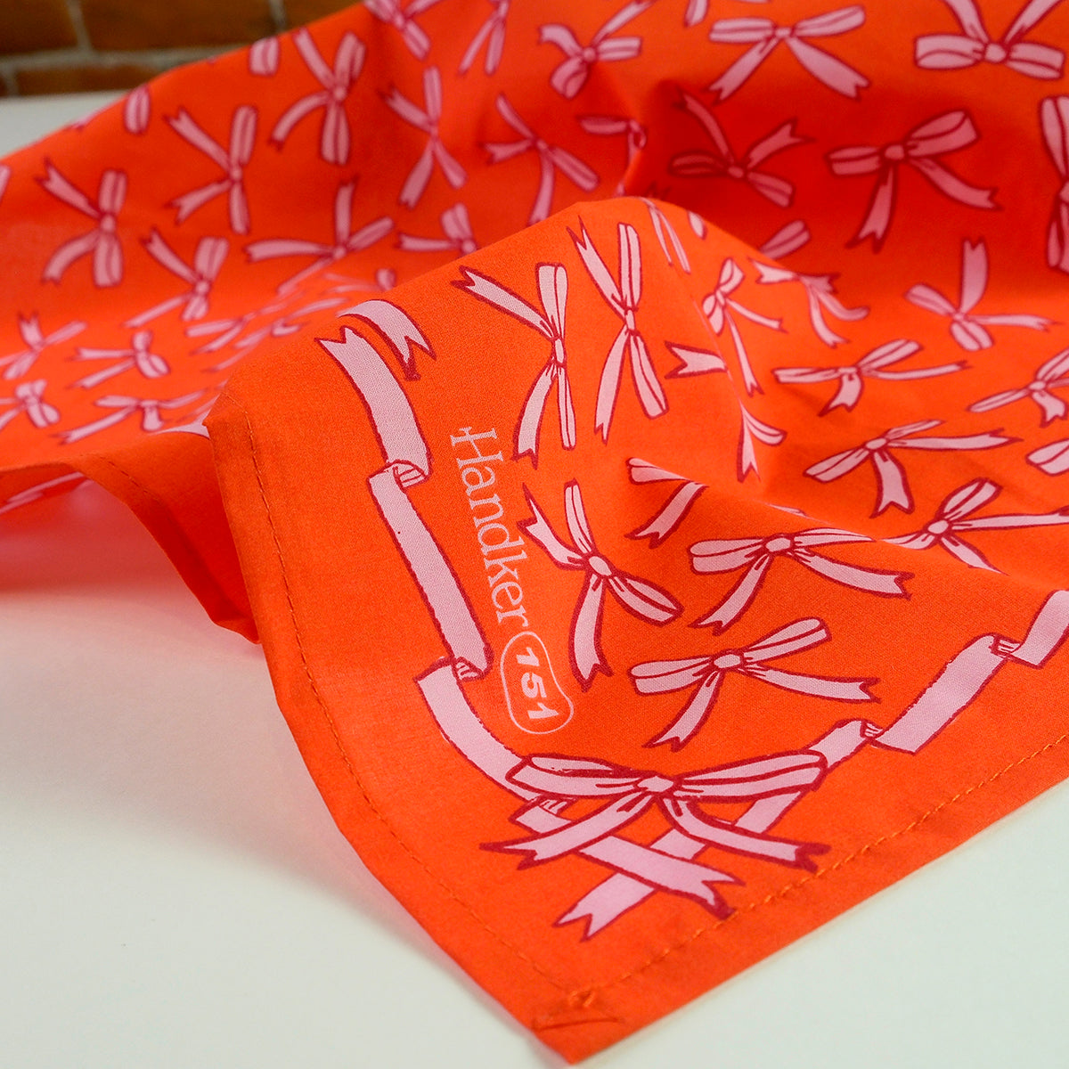 Close-up of No. 151 Beth bandana showing orange-red fabric with pink bows and 'Handker 151' corner print