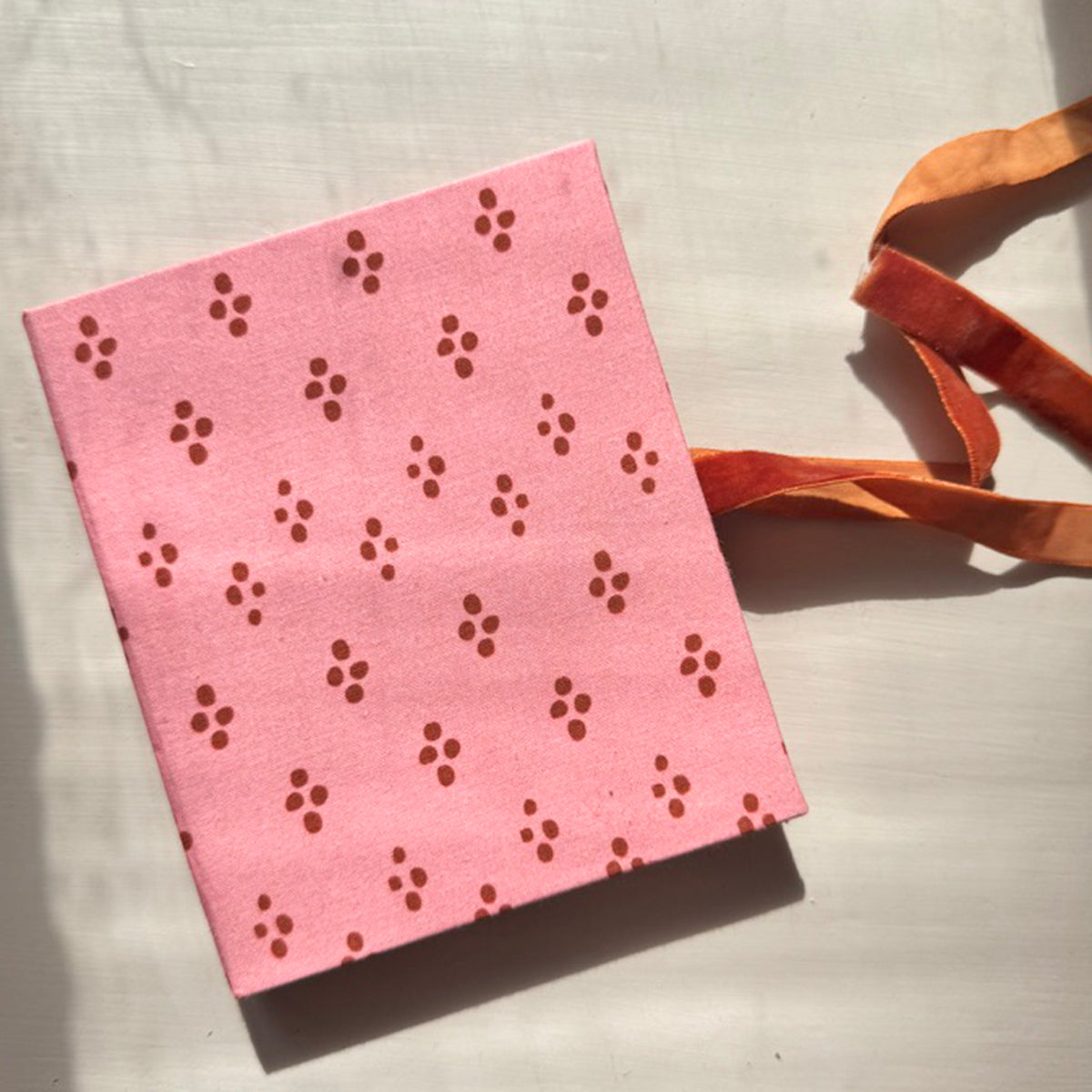 Pink notebook with punched flower-pattern cover casting shadow; orange ribbon beside it