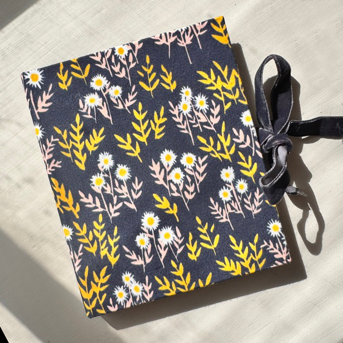 Fabric-covered notebook with navy background, white and yellow floral pattern and a gray ribbon tie.