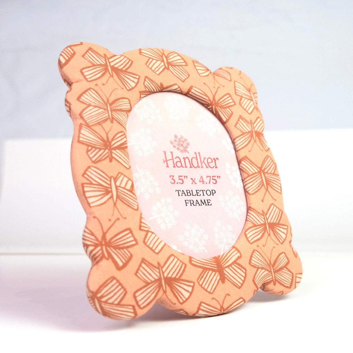 Angled front view of Sally fabric-covered 6x7 tabletop frame with peachy pink butterfly pattern and 3.5 x 4.75 in photo opening on white background