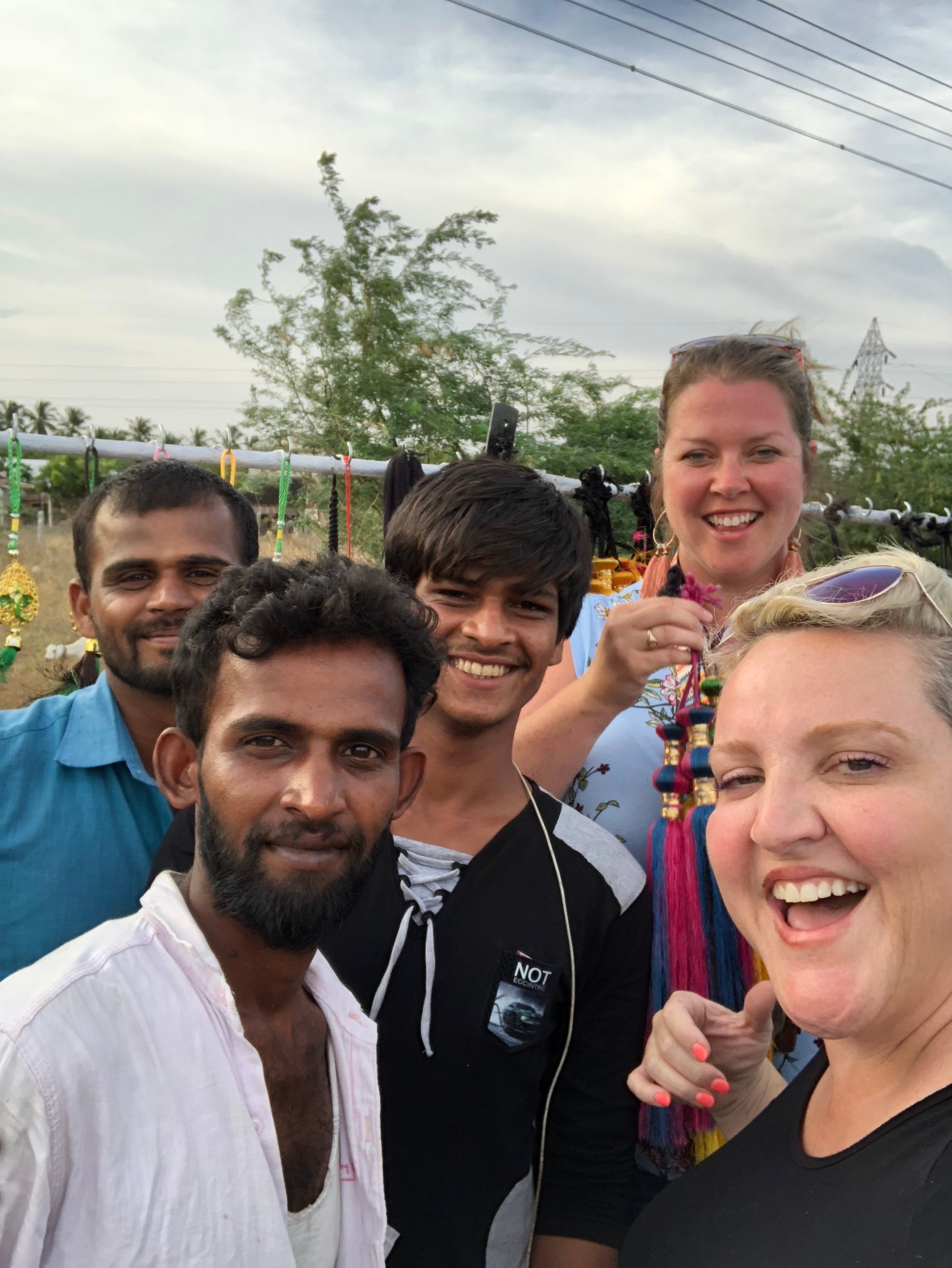Two women and three men smiling for a selfie at an outdoor Indian market, showing colorful hanging tassel ornaments