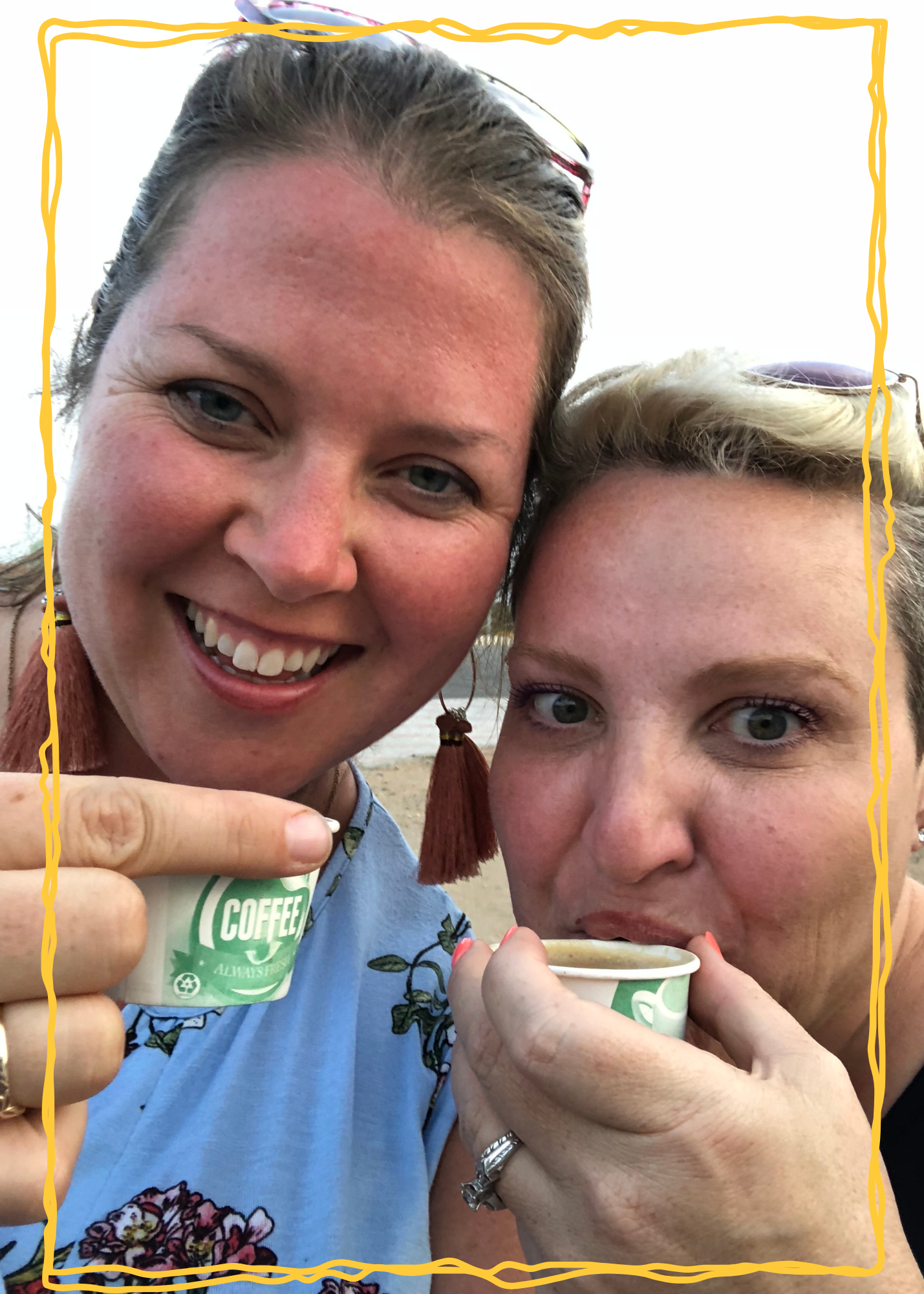 Two women smiling in a close-up selfie holding small paper cups labeled COFFEE on a beach