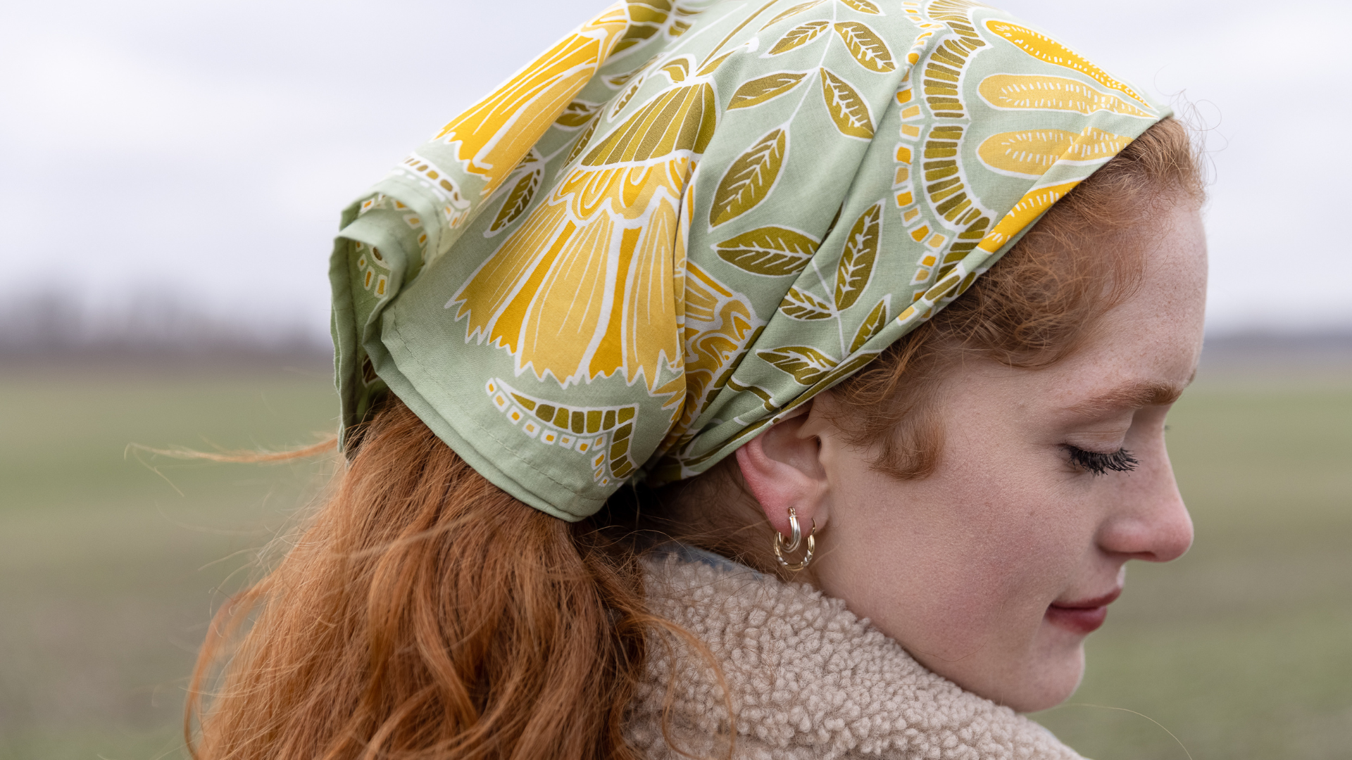 Profile of a woman wearing a green and yellow floral bandana tied as a headscarf over red hair in an open field