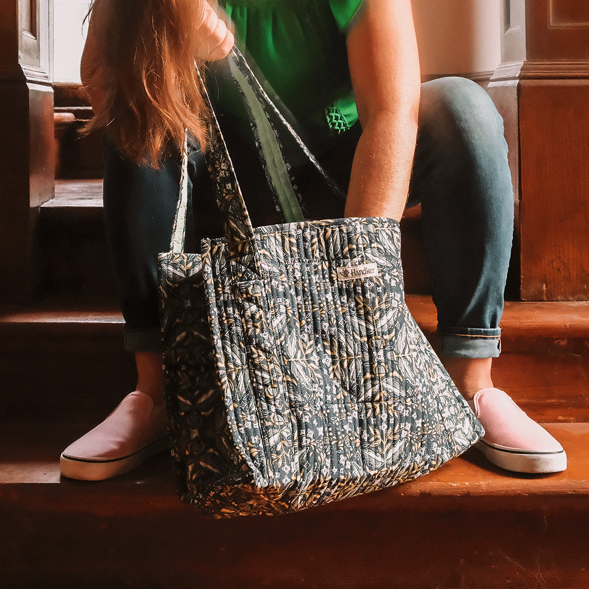 Quilted patterned tote bag with chain straps resting on wooden stairs beside a person in jeans and pink flats