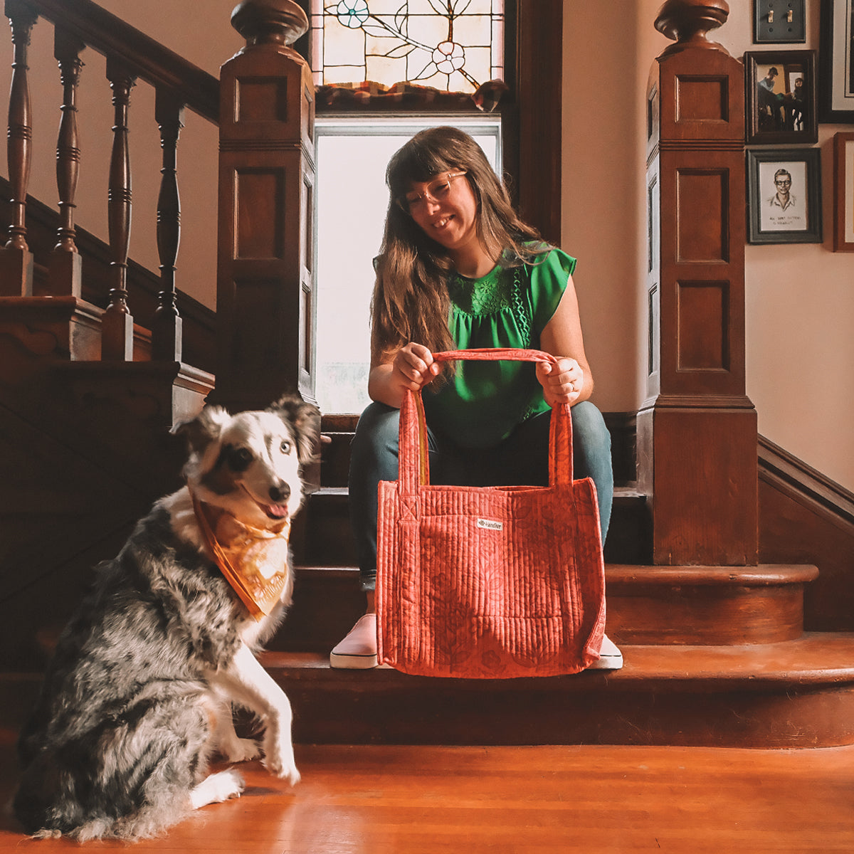 Woman in green dress on wooden staircase holding a large copper tote bag, collie dog seated beside her.