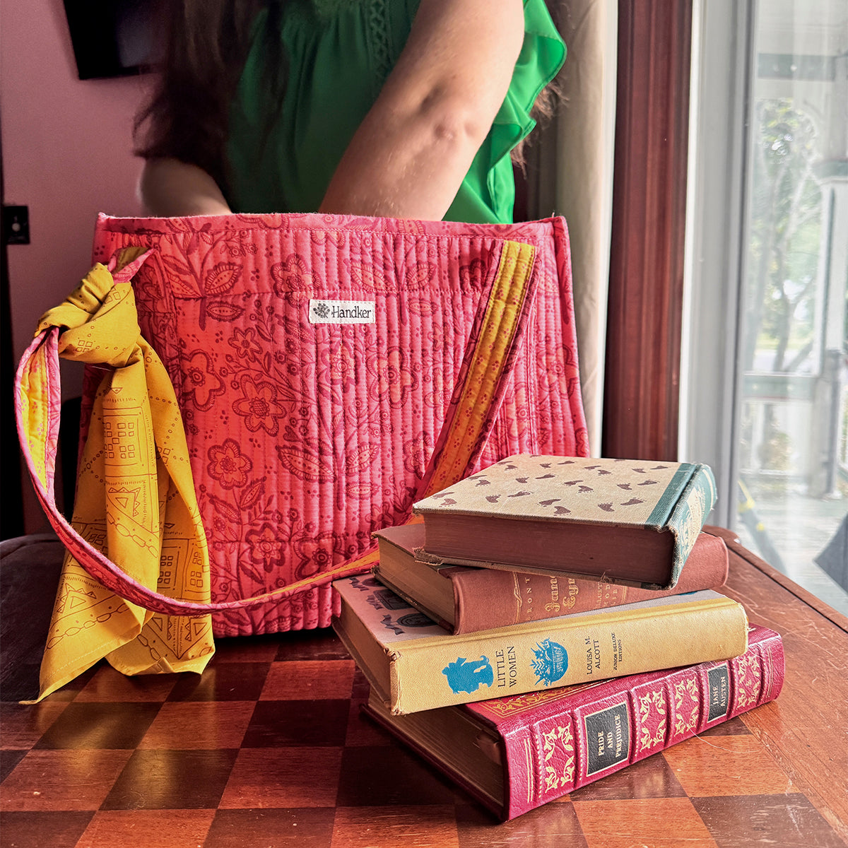 Pink quilted handbag with a yellow strap on a wooden surface next to stacked books.