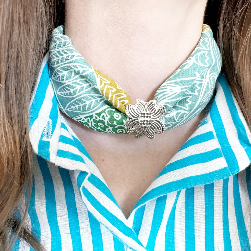 Green and aqua patterned silk neck scarf tied with a silver floral clasp at center front