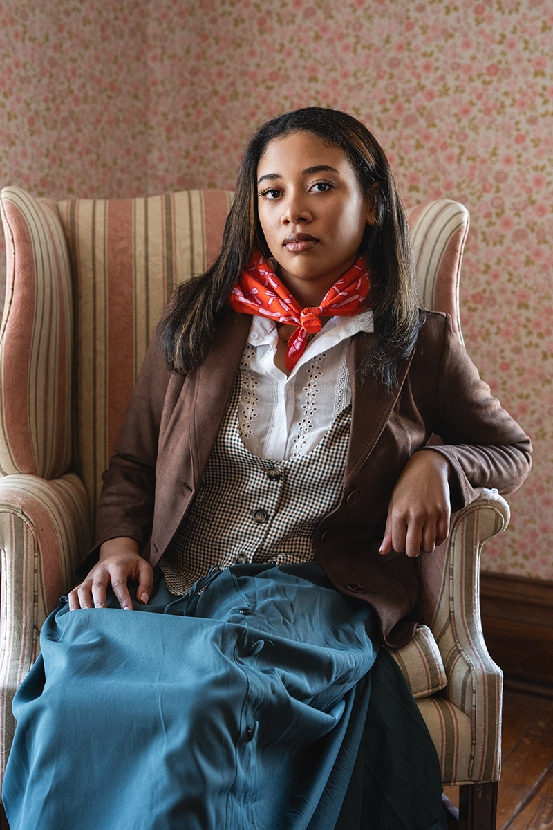Woman sitting in a chair wearing a brown blazer, white shirt, orange-red bandana, and blue skirt against a floral wallpapered wall.