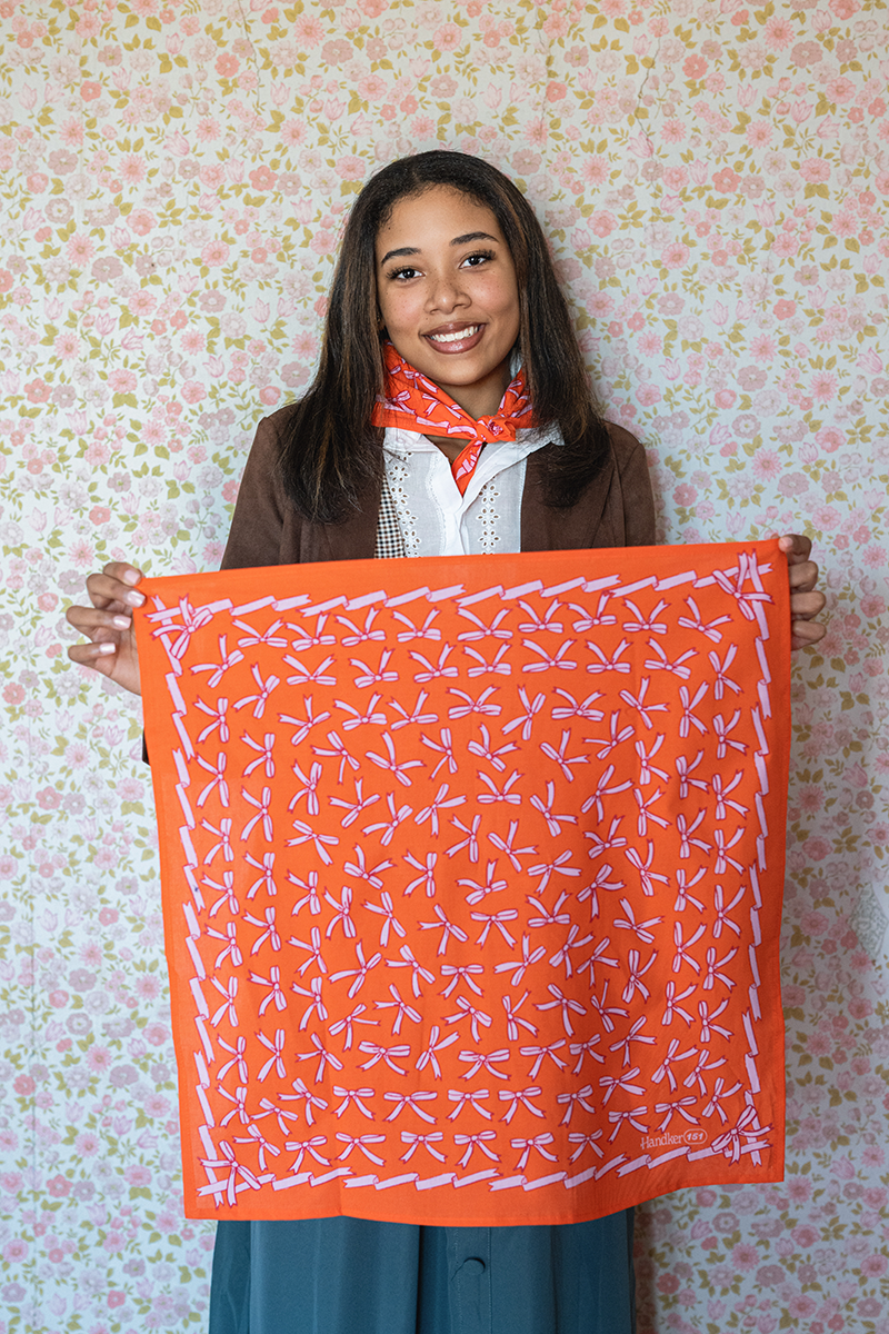 Woman holding an orange bandana with a pink bow pattern against a floral wallpaper background