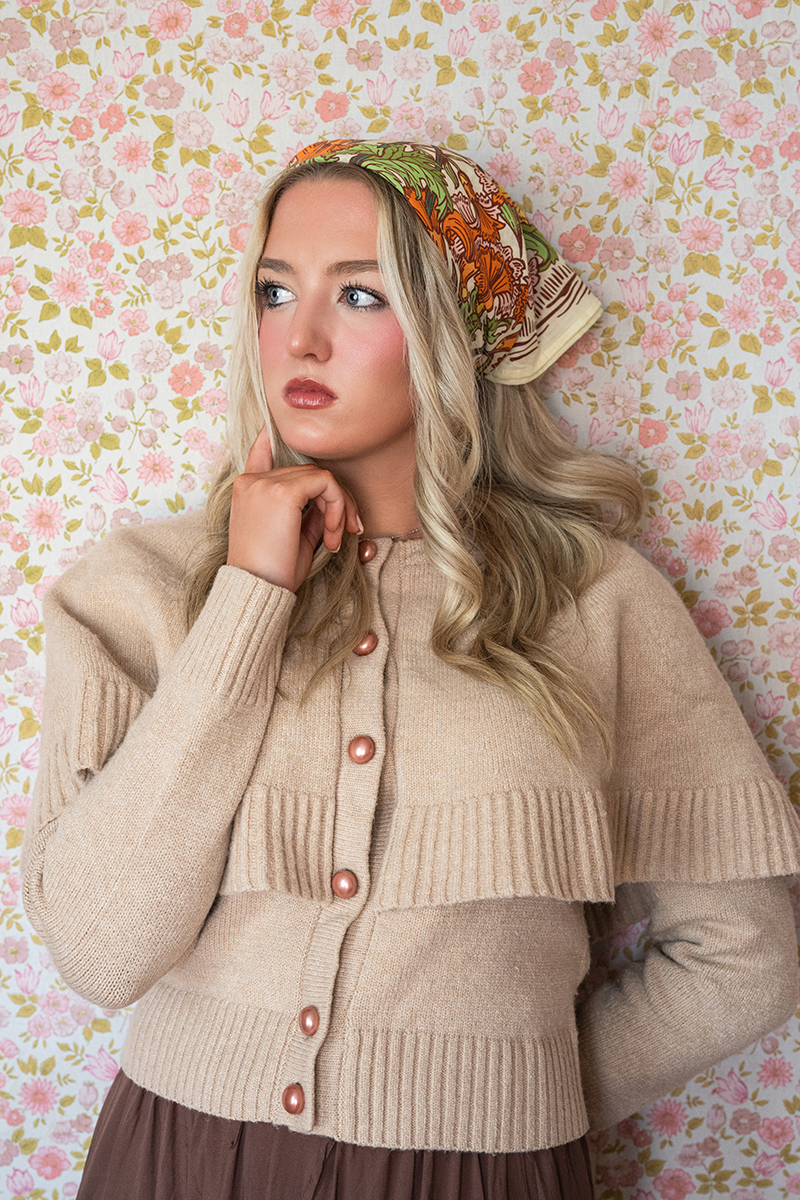 Woman wearing a beige cardigan and cream bandana with orange and green floral pattern against a floral wallpaper background