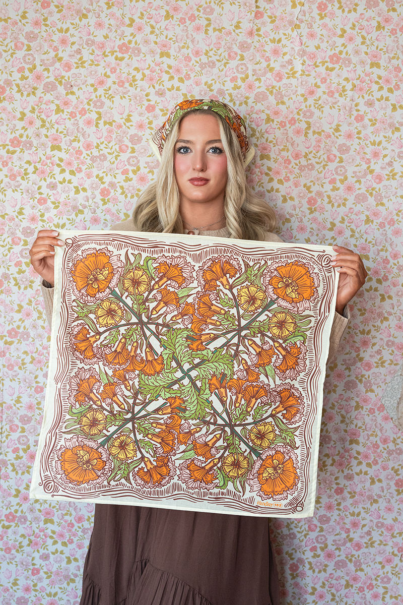 Woman holding a cream, orange, yellow, and green floral patterned bandana against a floral wallpaper background