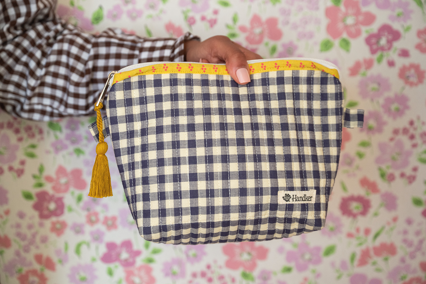 Hand holding a blue and white checkered pouch with a yellow zipper against a floral background