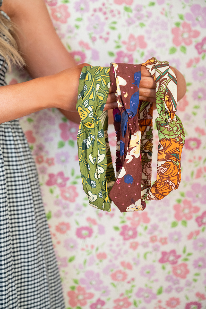 Close-up of three Hannah Cotton Headbands held by hand showcasing printed cotton fabric and elastic back with pink floral background