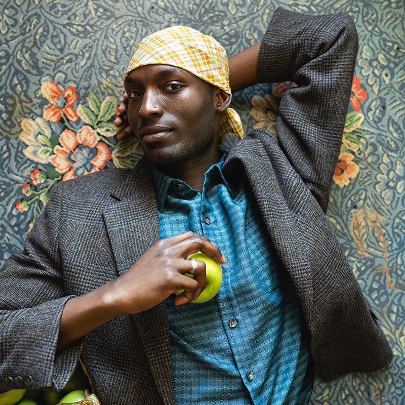Man reclining on patterned floral rug wearing yellow plaid bandana and brown checked blazer while holding a green apple