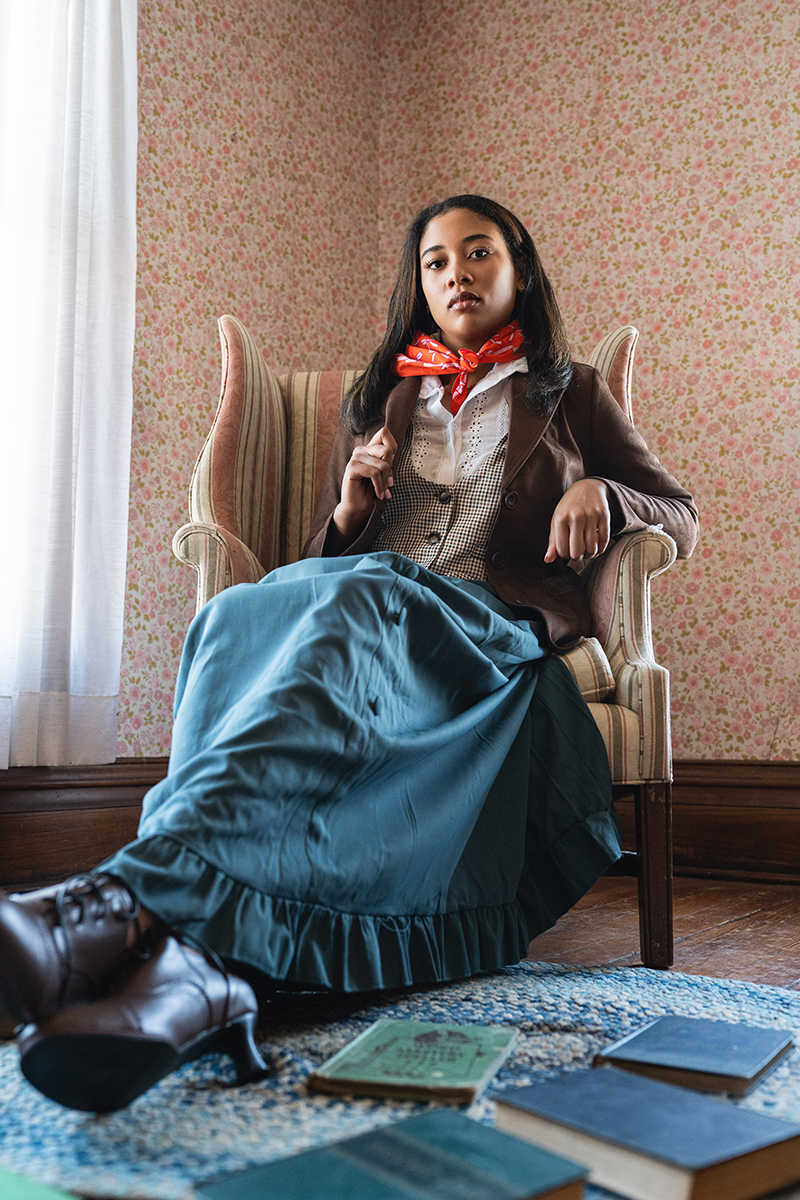 Woman seated in an armchair wearing an orange bandana tied at her neck, brown jacket and teal skirt in a floral room