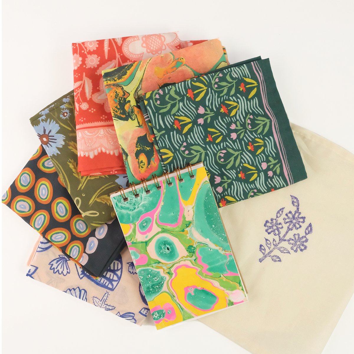 Colorful patterned fabric squares with a marbled spiral notebook on a cream tote stamped with purple flowers.