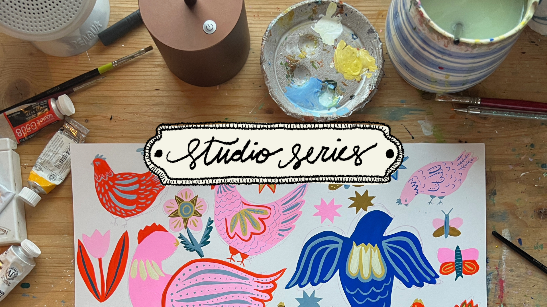 Wood desk with paintbrushes, palette and a colorful folk-style illustration of birds and flowers labeled 'studio series'.