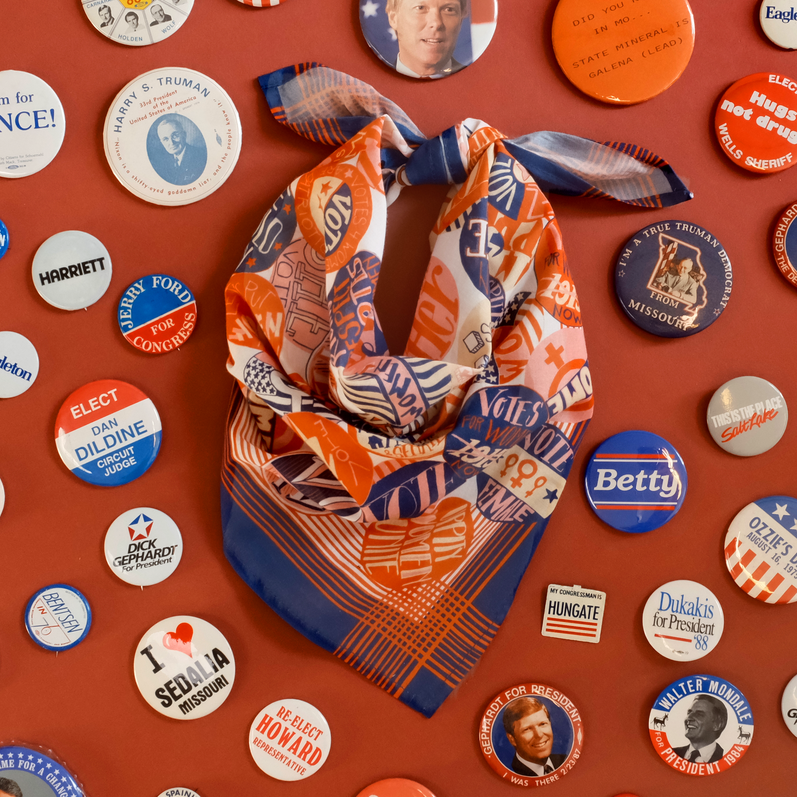 Assorted political campaign buttons and an orange patterned scarf, including buttons reading 'Biden', 'Biden Harris', 'Beto', and 'I Voted'.