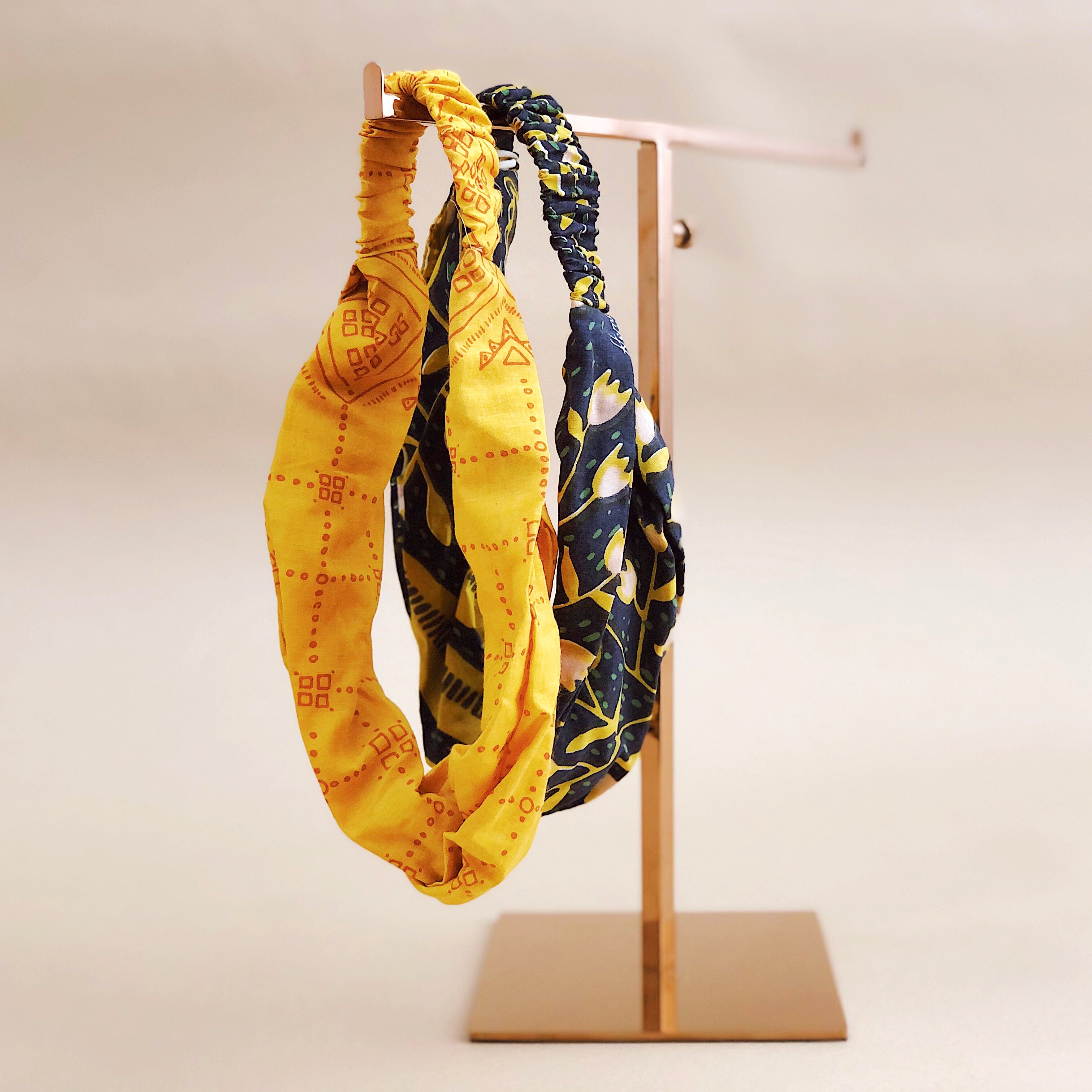 Front view of two knotted fabric headbands showcasing ruched elastic backs and yellow tulip and navy floral prints on a gold display stand