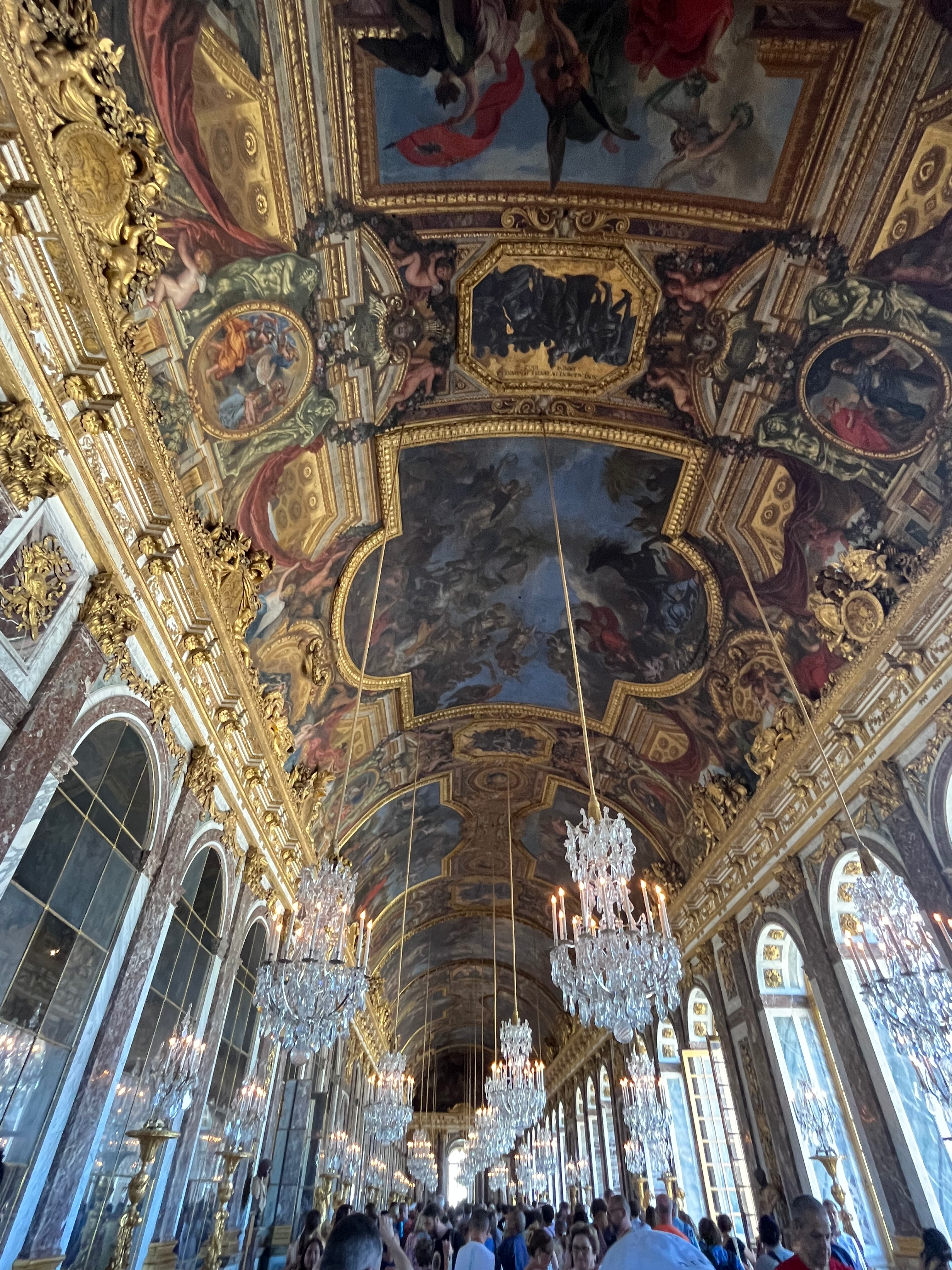 Visitors inside the Hall of Mirrors at the Palace of Versailles in Paris, beneath ornate painted ceilings and crystal chandeliers