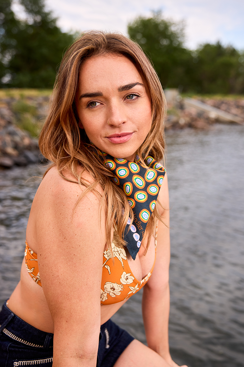 Woman wearing No. 142 Lorena bandana (orange, gold, and pink on navy) tied at her neck while seated on a rocky lakeshore