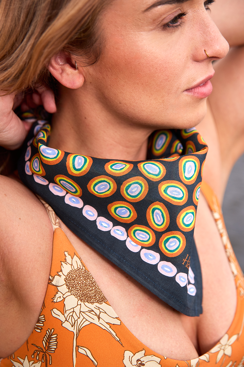 Close-up of No. 142 Lorena bandana showcasing concentric orange, gold, and pink circles on a navy blue background tied around a model's neck