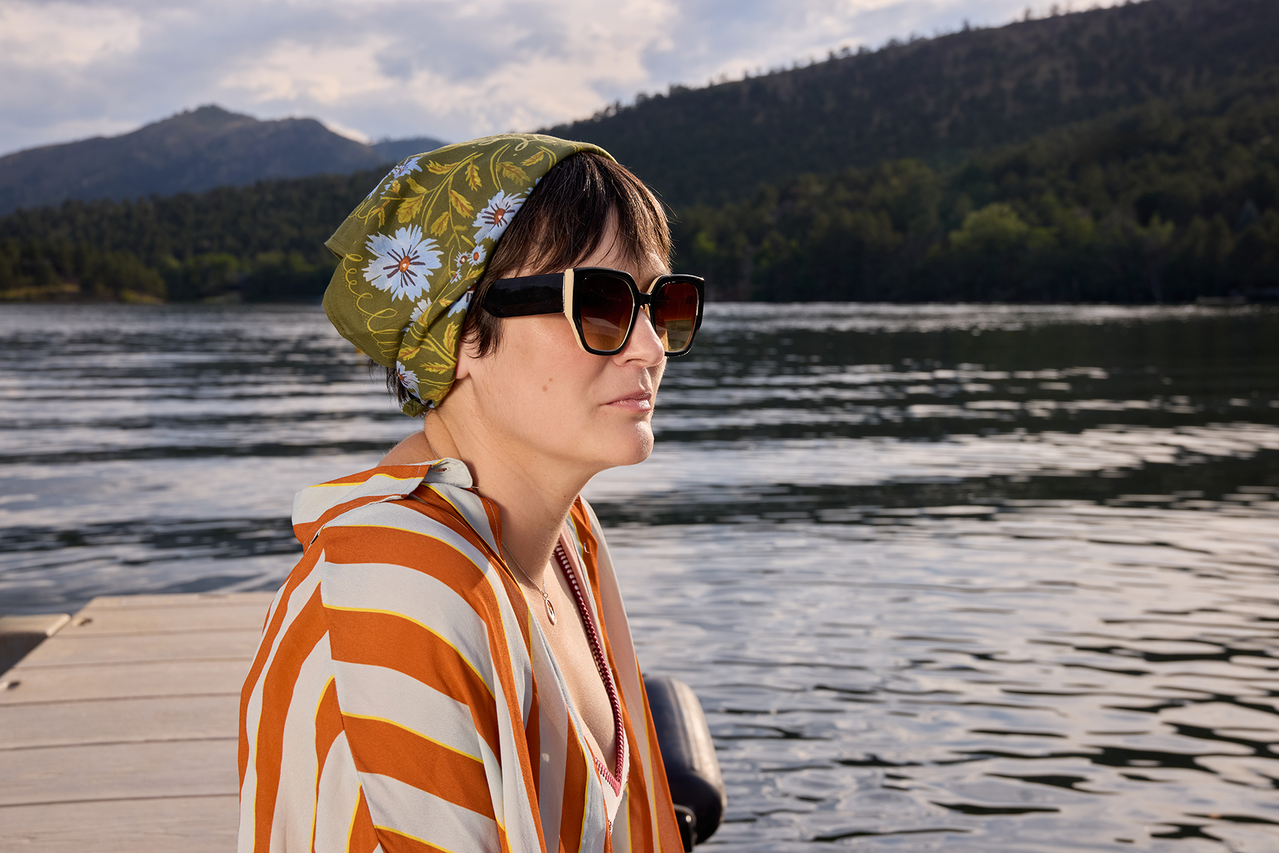 Woman wearing olive green bandana with periwinkle floral print tied as headscarf, sitting on a dock by a mountain lake