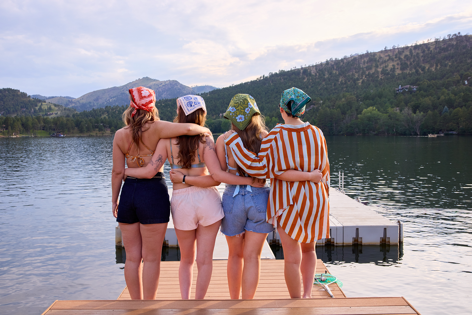 Four people standing arm-in-arm on a wooden dock by a mountain lake, each wearing a patterned bandana