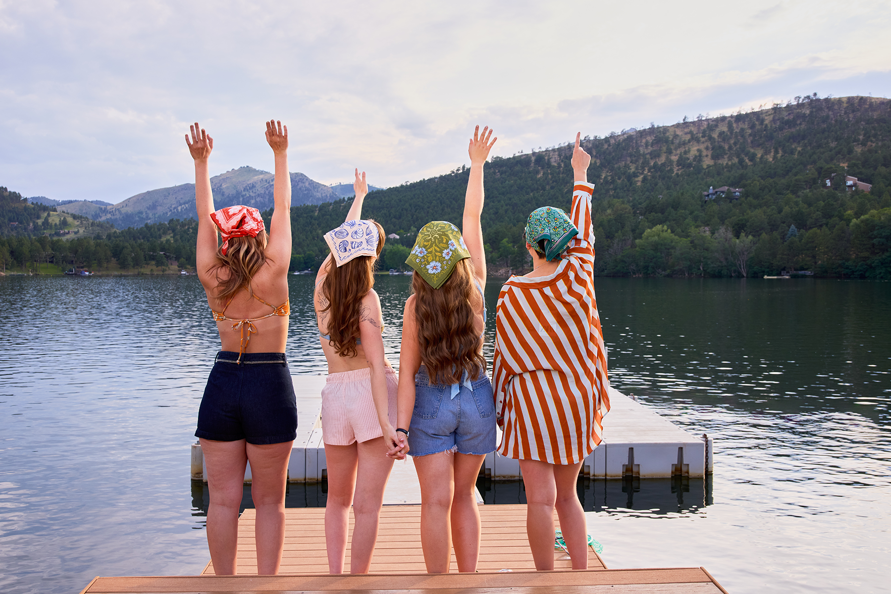 Four women on a lakeside dock raising their arms, one wearing the No. 143 Clara olive-green periwinkle bandana
