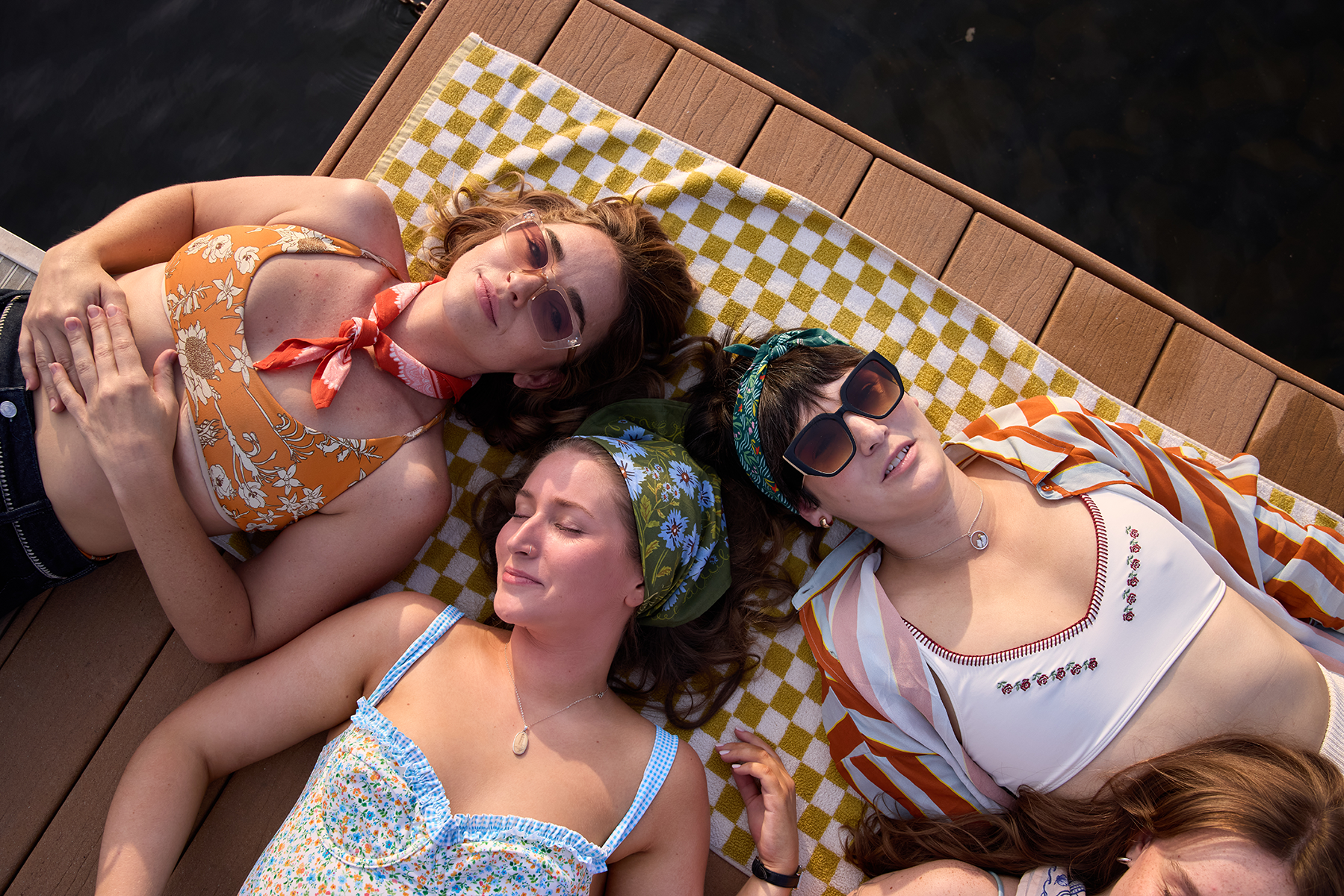Three women lying on a wooden dock by the water wearing colorful bandanas as headscarves and neck scarves on a yellow check towel
