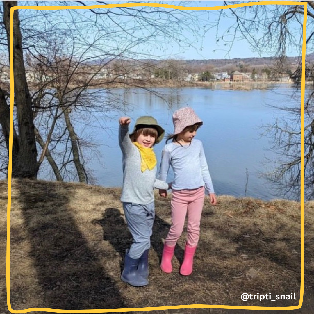 Two children holding hands by a lake; one with a yellow bandana and blue boots, the other with a pink hat and pink boots