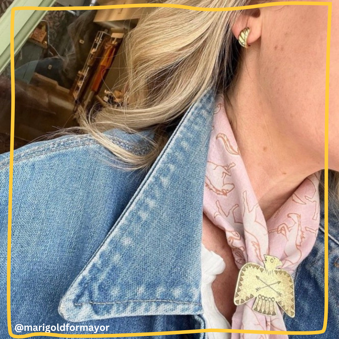 Woman wearing a pale pink bandana with tan horse print tied at her neck with a gold eagle scarf slide, styled with a light denim jacket