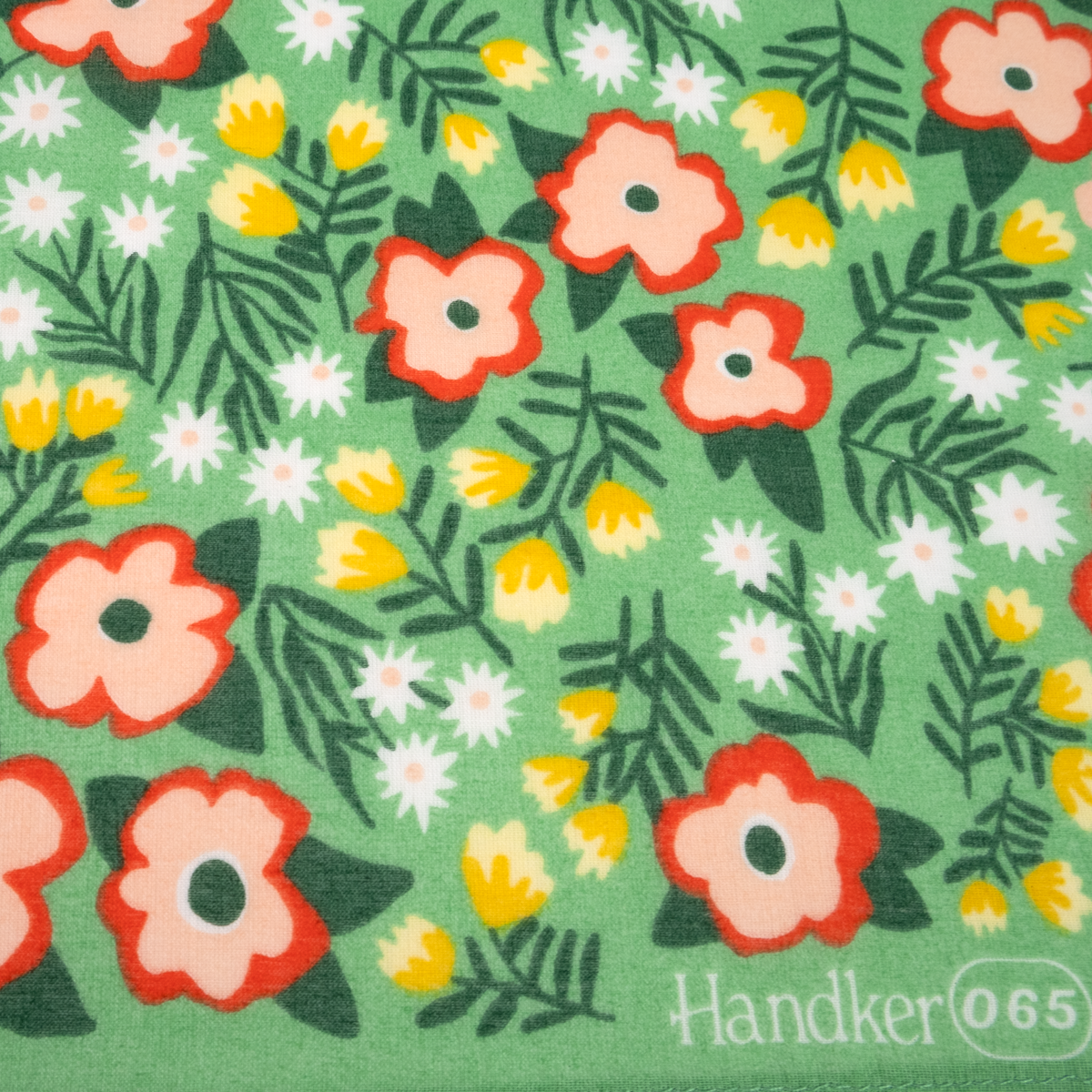 A close-up of a green bandana with a retro floral pattern featuring pink, red, yellow, and white flowers with dark green leaves. The text 'Handker(065)' is printed in white in the bottom right corner.