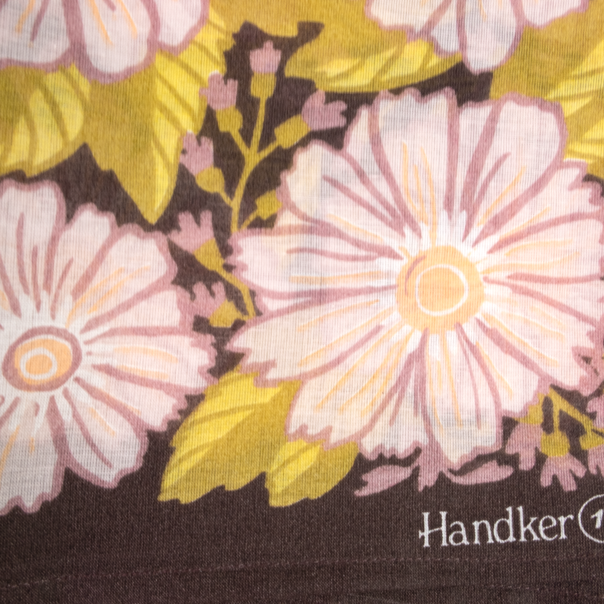A close-up of a bandana featuring large pale pink flowers with yellow-green leaves on a dark brown background. The brand name 'Handker' is partially visible in white text in the bottom right corner.
