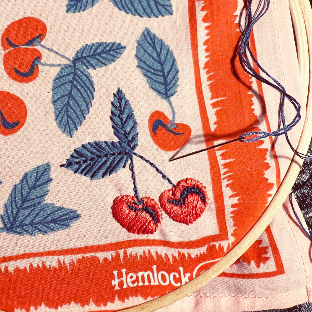Hand-embroidered bandana with red cherries and blue leaves in a wooden hoop, Hemlock printed on the border